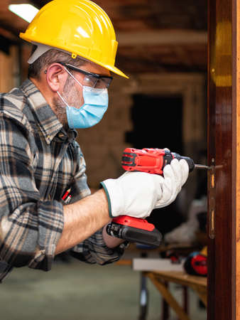 Carpenter worker at work repairs and installs a room door, wear the surgical mask to prevent Coronavirus infection. Preventing Pandemic Covid-19 at the workplace. Carpentry.の写真素材