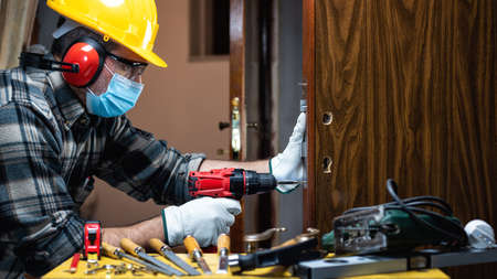 Carpenter worker at work repair and install a room door lock, wear the surgical mask to prevent Coronavirus infection. Preventing Pandemic Covid-19 at the workplace. Carpentry.の写真素材