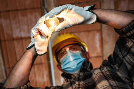 Electrician worker at work replaces the light bulb protected by helmet, safety goggles and glovesの写真素材