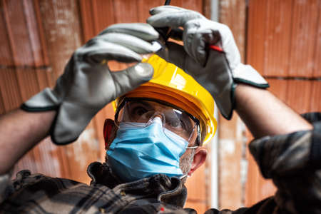 Electrician worker at work replaces the light bulb protected by helmet, safety goggles and gloves. Wear the surgical mask to prevent the spread of virus. Construction industry.の写真素材