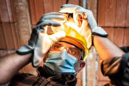 Electrician worker at work replaces the light bulb protected by helmet, safety goggles and gloves. Wear the surgical mask to prevent the spread of virusの写真素材