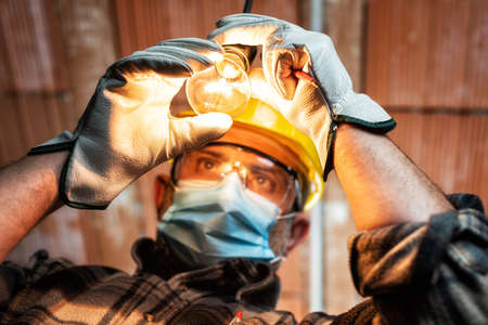Electrician worker at work replaces the light bulb protected by helmet, safety goggles and gloves. Wear the surgical mask to prevent the spread of virusの写真素材