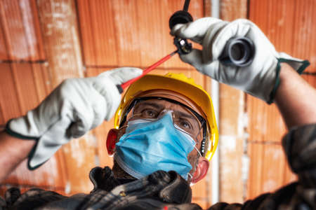 Electrician worker at work replaces the lamp holder protected by helmet, safety goggles and gloves; wear the surgical mask to prevent the spread of virusの写真素材