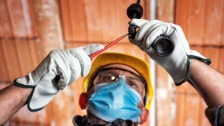 Electrician worker at work replaces the lamp holder protected by helmet, safety goggles and gloves; wear the surgical mask to prevent the spread virusの写真素材
