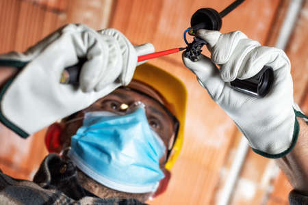 Electrician worker at work replaces the lamp holder protected by helmet, safety goggles and gloves; wear the surgical mask to prevent the virusの写真素材