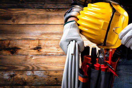 Electrician worker on vintage wooden background; holds the roll of electric cable, helmet and protective goggles in his hand. Construction industry, electrical system.の写真素材