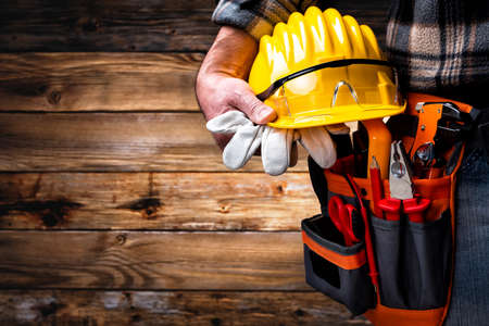 Electrician worker on vintage wooden background; holds helmet, gloves and goggles in hand. Construction industry, electrical system.の写真素材