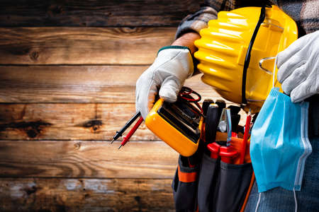 Electrician worker on vintage wooden background; holds the tester, helmet, protective goggles and the surgical mask to prevent the spread of Coronavirus. Construction industry, work safety.の写真素材
