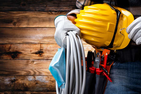 Electrician worker on vintage wooden background; holds the roll of electric cable, helmet, protective goggles and the surgical mask to prevent the spread of Coronavirus. Construction industry, work safety.の写真素材