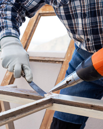 Carpenter using heat gun and wood scraper removes paint from old wooden window. Construction industry, carpentry. Restoration.の写真素材