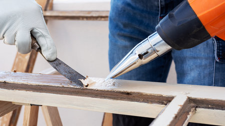 Carpenter using heat gun and wood scraper removes paint from old wooden window. Construction industry, carpentry. Restoration.の写真素材