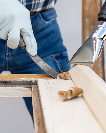 Carpenter using heat gun and wood scraper removes paint from old wooden window. Construction industry, carpentry. Restoration.の写真素材