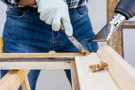 Carpenter using heat gun and wood scraper removes paint from old wooden window. Construction industry, carpentry. Restoration.の写真素材