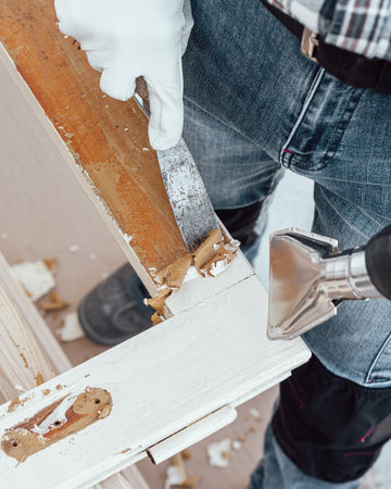 Carpenter using heat gun and wood scraper removes paint from old wooden window. Construction industry, carpentry. Restoration.の写真素材