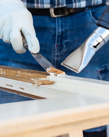 Carpenter using heat gun and wood scraper removes paint from old wooden window. Construction industry, carpentry. Restoration.の写真素材