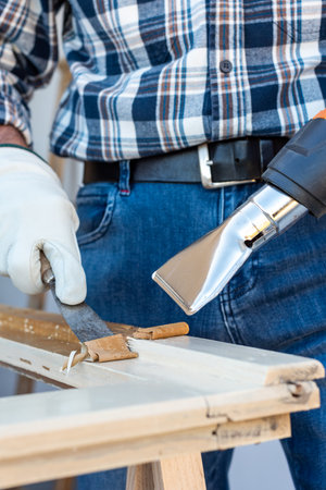 Carpenter using heat gun and wood scraper removes paint from old wooden window. Construction industry, carpentry. Restoration.の写真素材