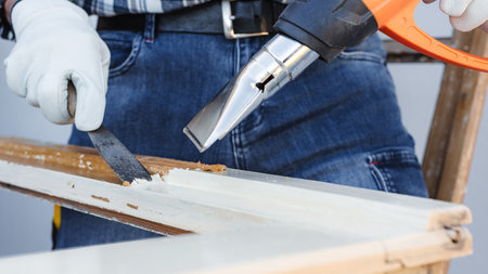 Carpenter using heat gun and wood scraper removes paint from old wooden window. Construction industry, carpentry. Restoration.の写真素材