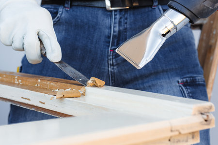 Carpenter using heat gun and wood scraper removes paint from old wooden window. Construction industry, carpentry. Restoration.の写真素材