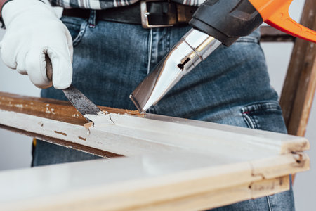 Carpenter using heat gun and wood scraper removes paint from old wooden window. Construction industry, carpentry. Restoration.の写真素材