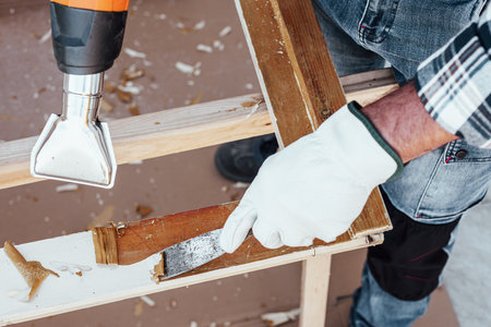Carpenter using heat gun and wood scraper removes paint from old wooden window. Construction industry, carpentry. Restoration.の写真素材