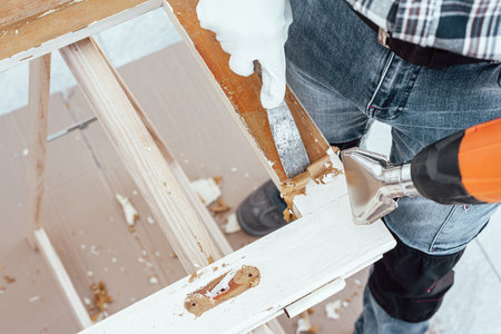 Carpenter using heat gun and wood scraper removes paint from old wooden window. Construction industry, carpentry. Restoration.の写真素材