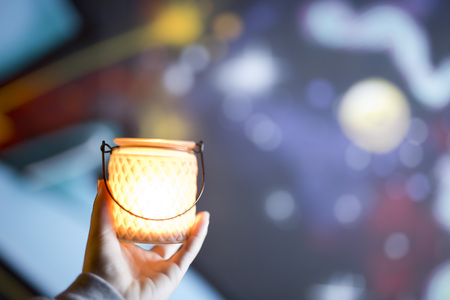 Container glass in the hand of a young woman, with a candle lit inside. Horizontal shot with colors out of focus background.の写真素材
