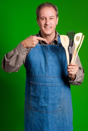 Man with an apron showing cooking tools against a green backgroundの写真素材