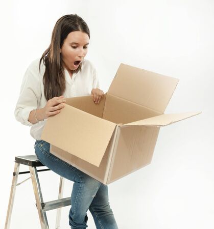 Young woman sitting on a ladder looking at an open cardboard box with a surprised expression