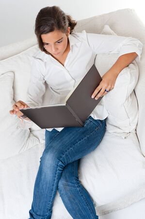 Young teenage girl reading a book sitting at home on a sofaの写真素材