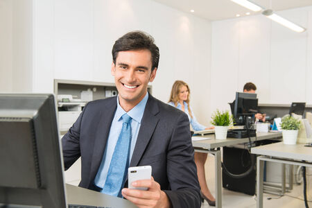 Young executive sitting at his desk, holding a Smartphoneの写真素材