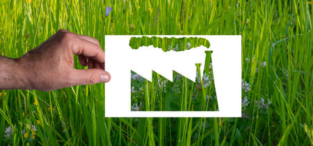 Green wash: A man's hand holds a blank sheet of paper with the silhouette of a factory cut out. Green grass background. Not everything "green" is ecologicalの写真素材