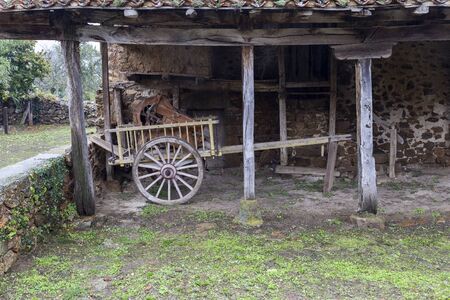 Vintage wooden carriage with old tillage machinery on top underneath a porch of a rural stone and wood houseの写真素材