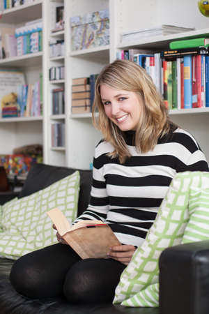 Young, attractive woman sits on the couch and reads a book at home  A bookshelf is at the background...