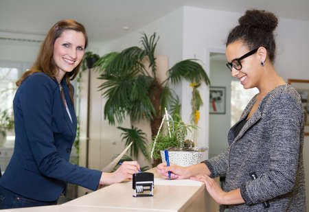 Smiling clerk (caucasian) is standing opposite to a customer (latin), who is filling out a document at a reception counter in the office.の写真素材