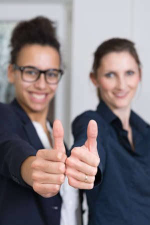 Two young business woman are standing side by side and are showing their thumbs up. Thumb is in focus, face is blurred.の写真素材