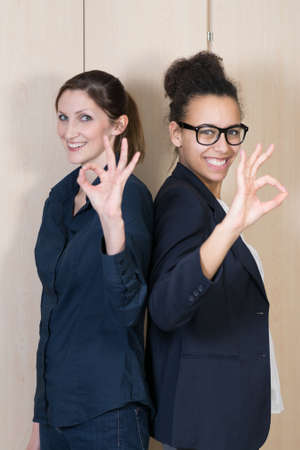 Two businesswomen are standing back to back and are forming a O with her fingers. Face is at focus, hand is blurred.の写真素材