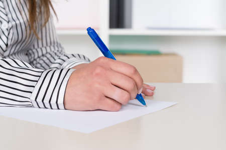 A businesswoman is writing with a blue pen at a piece of paper at the desk.