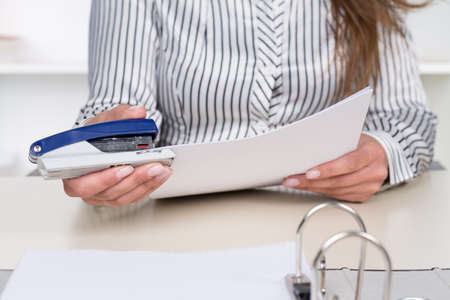 A businesswoman is stitching documents together at the desk in the office.