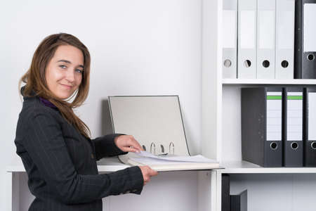 A young smiling businesswoman is browsing a file in front of a shelf in the office. The woman is looking to the camera.の写真素材