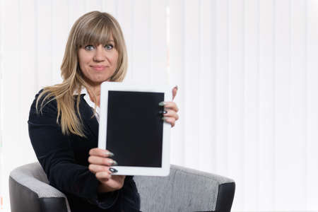 A businesswoman is showing a tablet while sitting on a chair. The woman is looking to the camera. Face is in focus, tablet is blurred.の写真素材