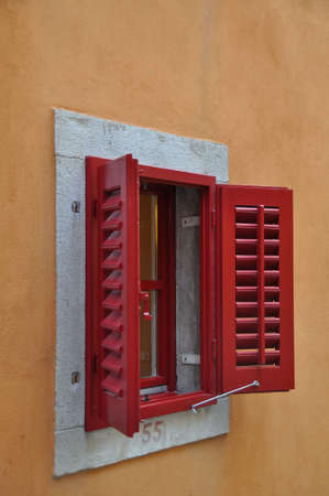 Red Window Shutters opened on a bright frame above a terracotta colored wall. The shutters unveil an...