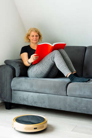 Vacuum cleaning robot with relaxed woman reading in the backgroundの写真素材
