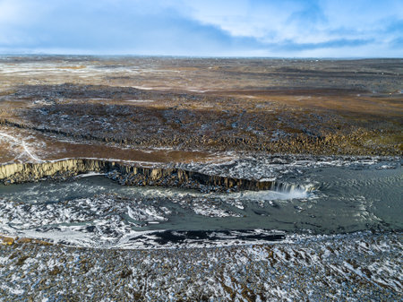Turquoise glacial river winds through a frozen Icelandic landscape, flowing toward the majestic Dettifoss waterfall, showcasing natures raw beauty and power from aboveの写真素材