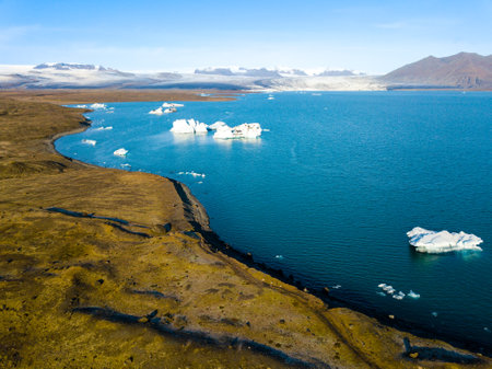 Aerial views capture the Icelandic glacier lagoon Jokulsarlon, featuring towering mountains and a tranquil lake beneath a bright blue skyの写真素材