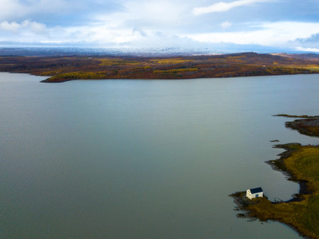 Small white house reflecting on the calm water of lake LagarfljÃ³t in EgilsstaÃ°ir, Iceland, on a cloudy day with snowy mountains in the backgroundの写真素材