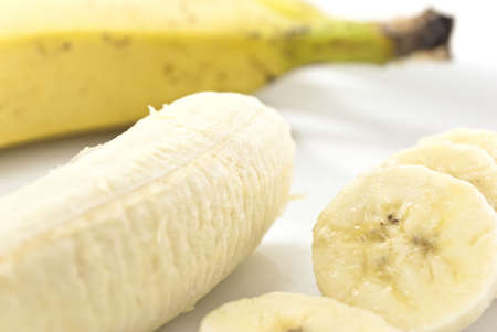 Close up shot of banana slices on a china plate, next to the remaining peeled banana portion, with an unpeeled banana in soft focus in the background. White background.の写真素材
