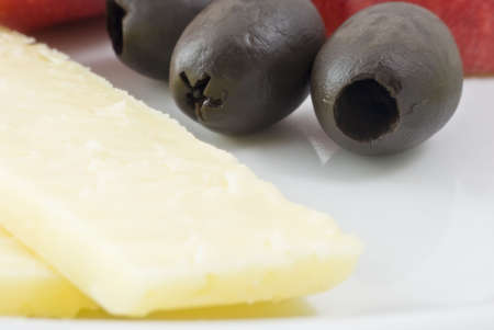 Macro shot of cheddar cheese and olives on a white plate.  Red apple just visible in the background.の写真素材