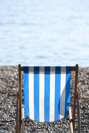 A solitary, empty, blue and white striped deckchair sitting on a pebble beach on a sunny day, with sea in soft focus in the background.の写真素材
