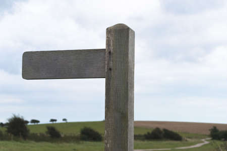 Natural wooden sign pointing West with copy space.  Hills of grass, a winding path, trees and cloudy blue sky in soft focus in the background.の写真素材