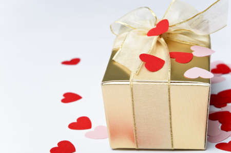 Close up of a gold gift box, tied and bowed with organza ribbon and scattered with pink and red paper heart confetti.  Background is off-white.の写真素材